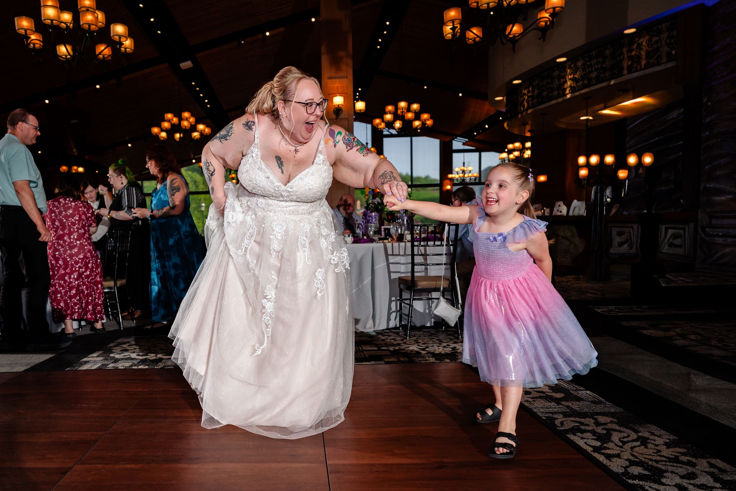 Bride in white gown joyfully dancing with young girl in pink dress on wedding reception dance floor.
