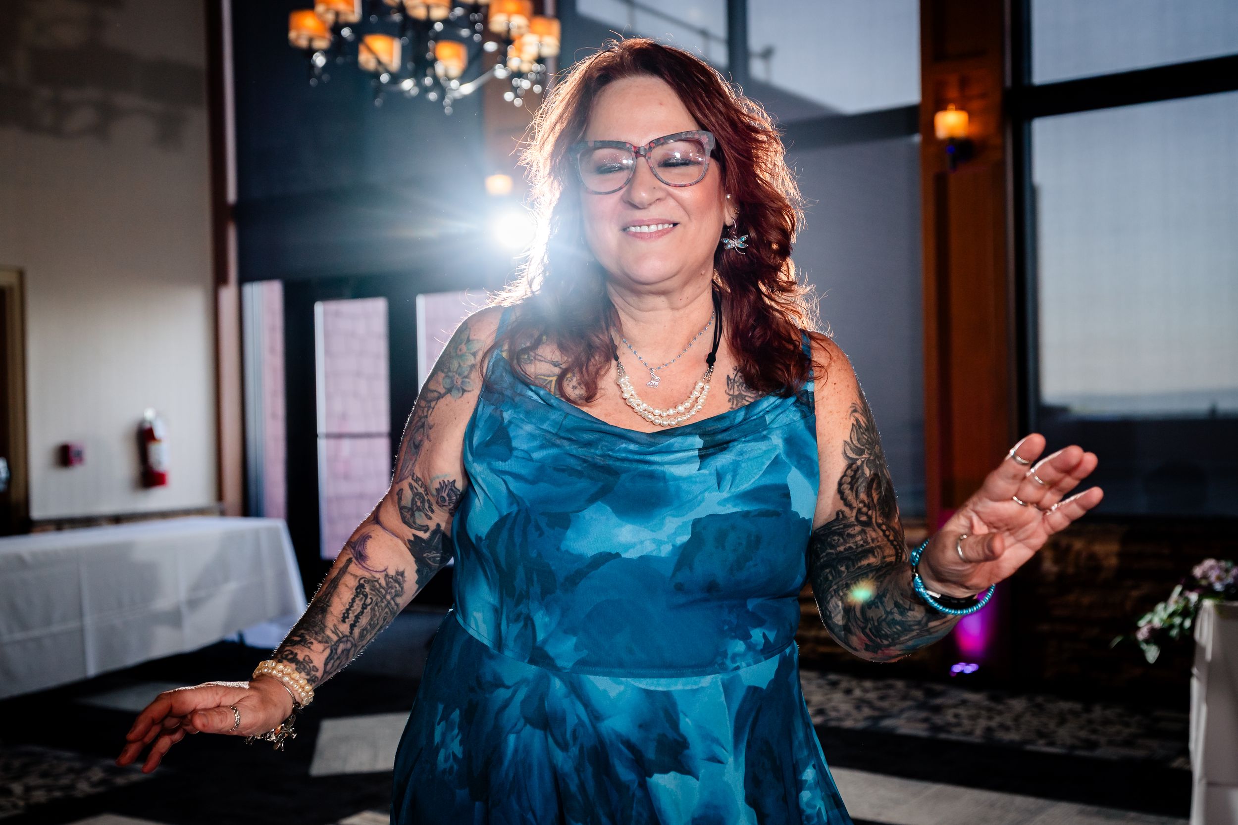 A woman with tattoos in a teal dress dances joyfully at a wedding reception with chandeliers behind her.