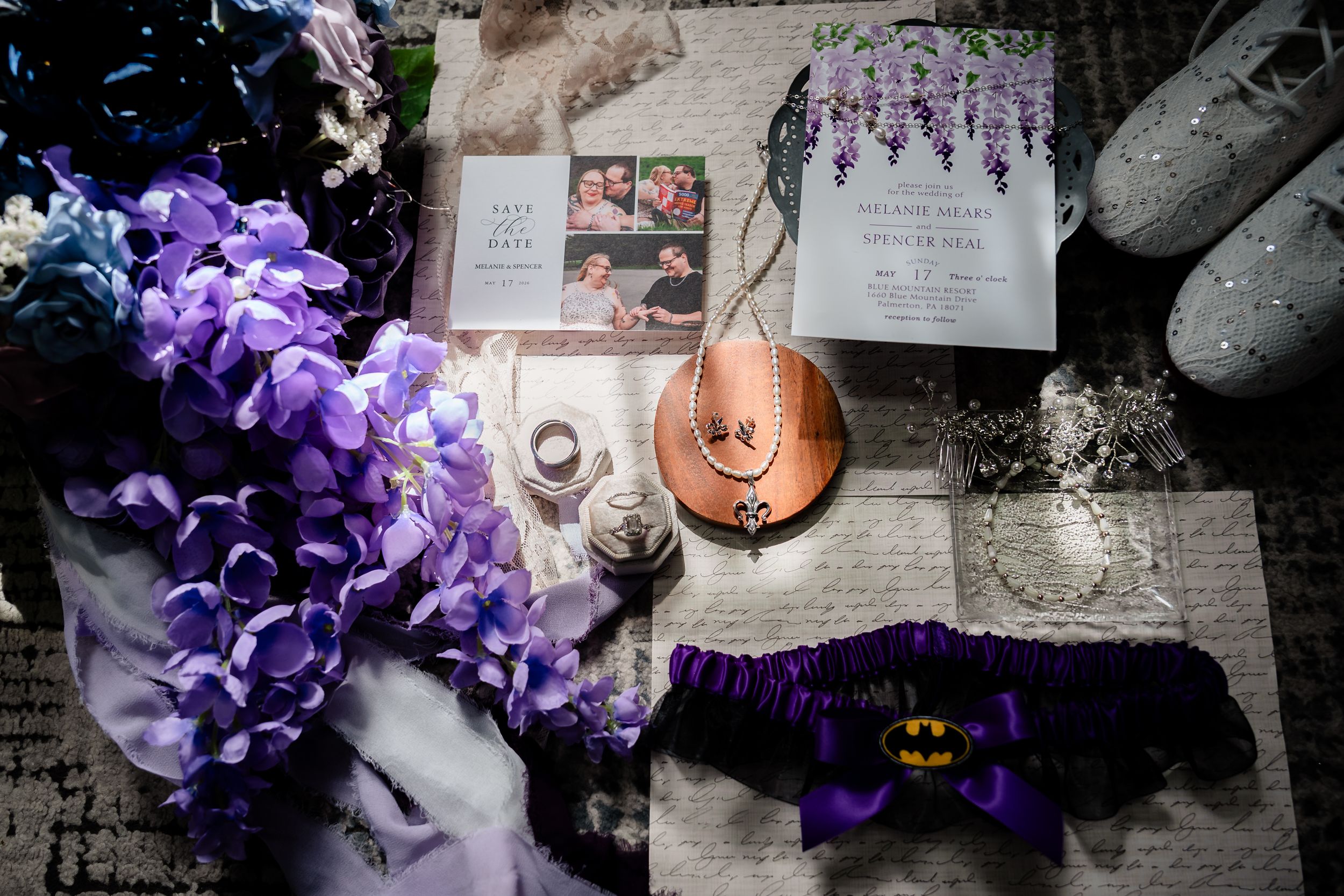 Purple floral wedding flat lay with Batman garter, invitations, rings, and bridal accessories on decorative paper.