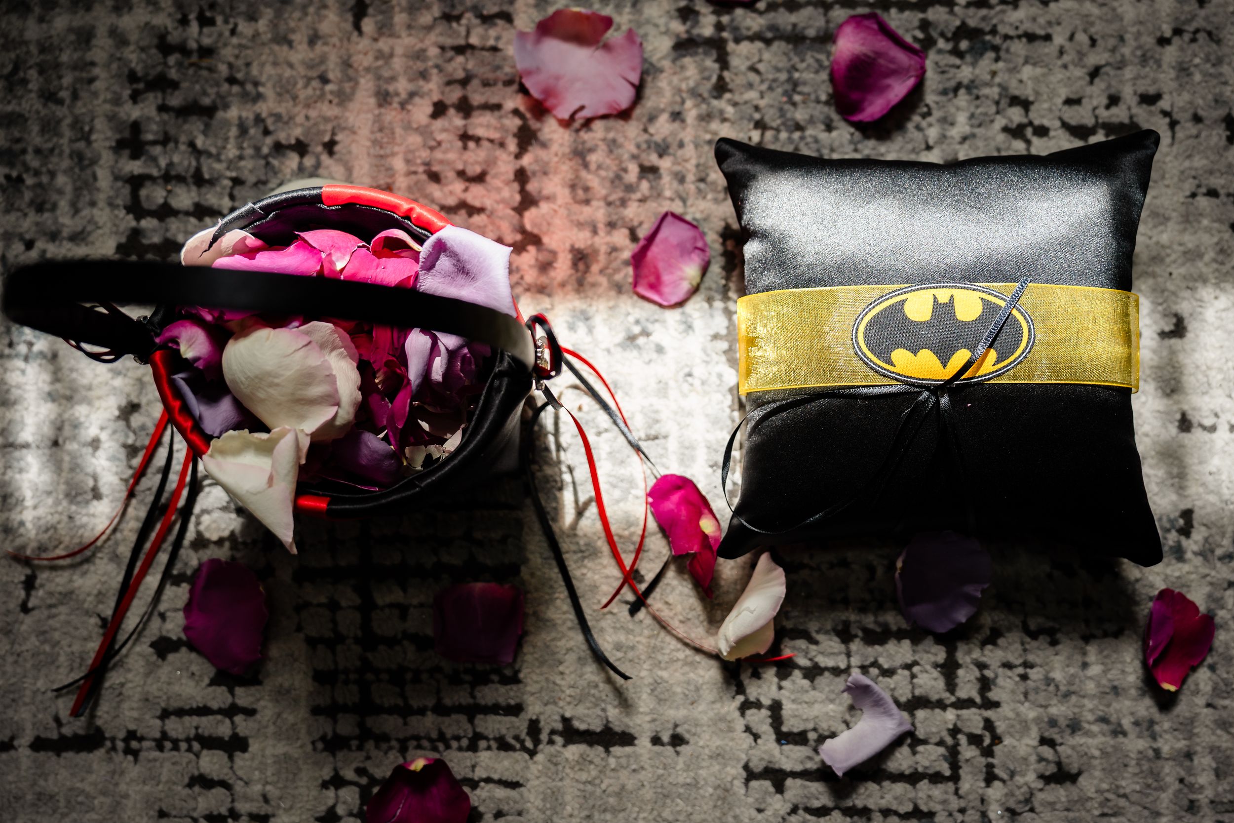 Batman pillow and floral accessories with rose petals scattered on a textured surface at a wedding.
