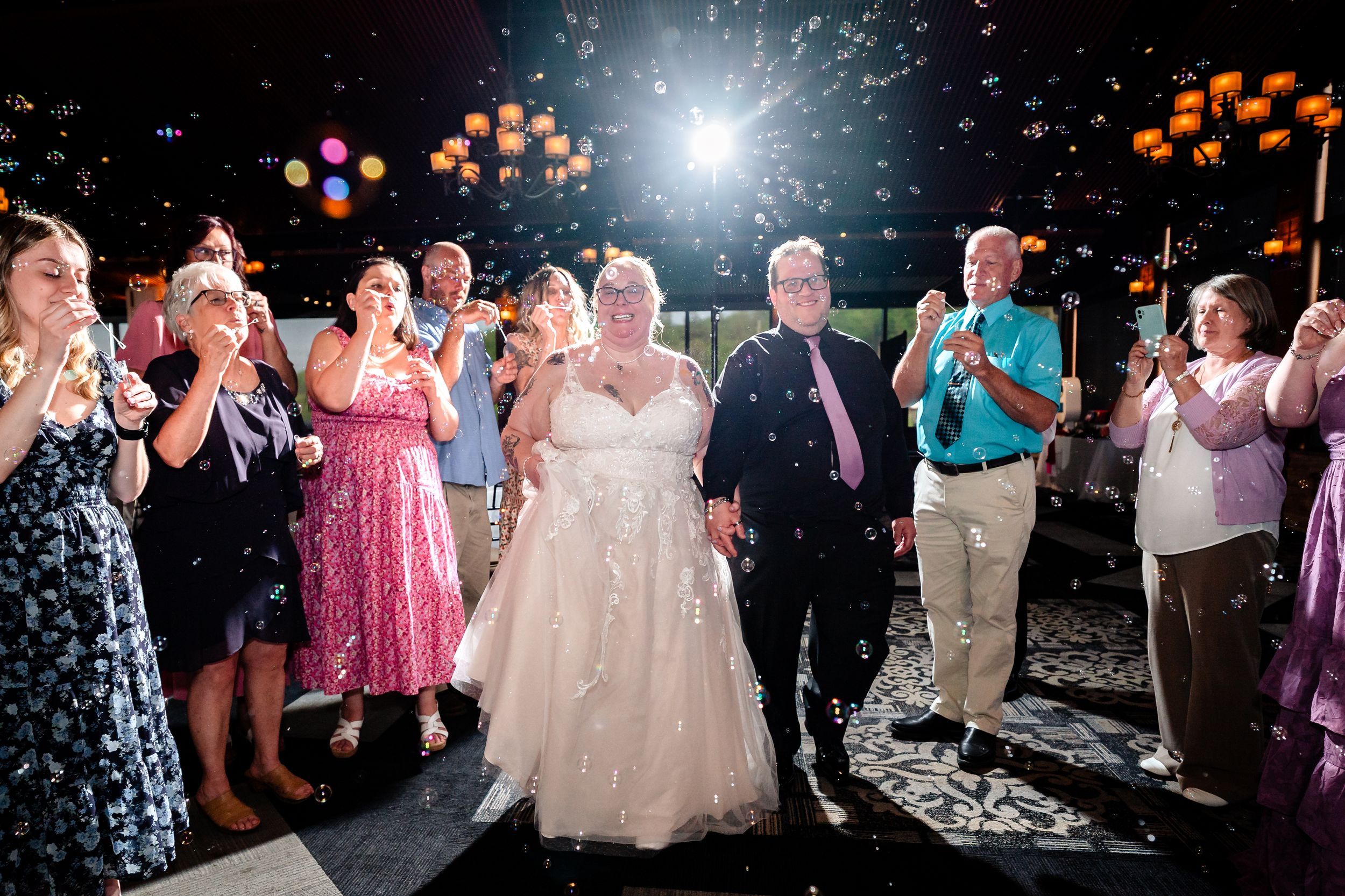Bride and groom walk through confetti celebration surrounded by cheering guests at outdoor wedding reception at night.
