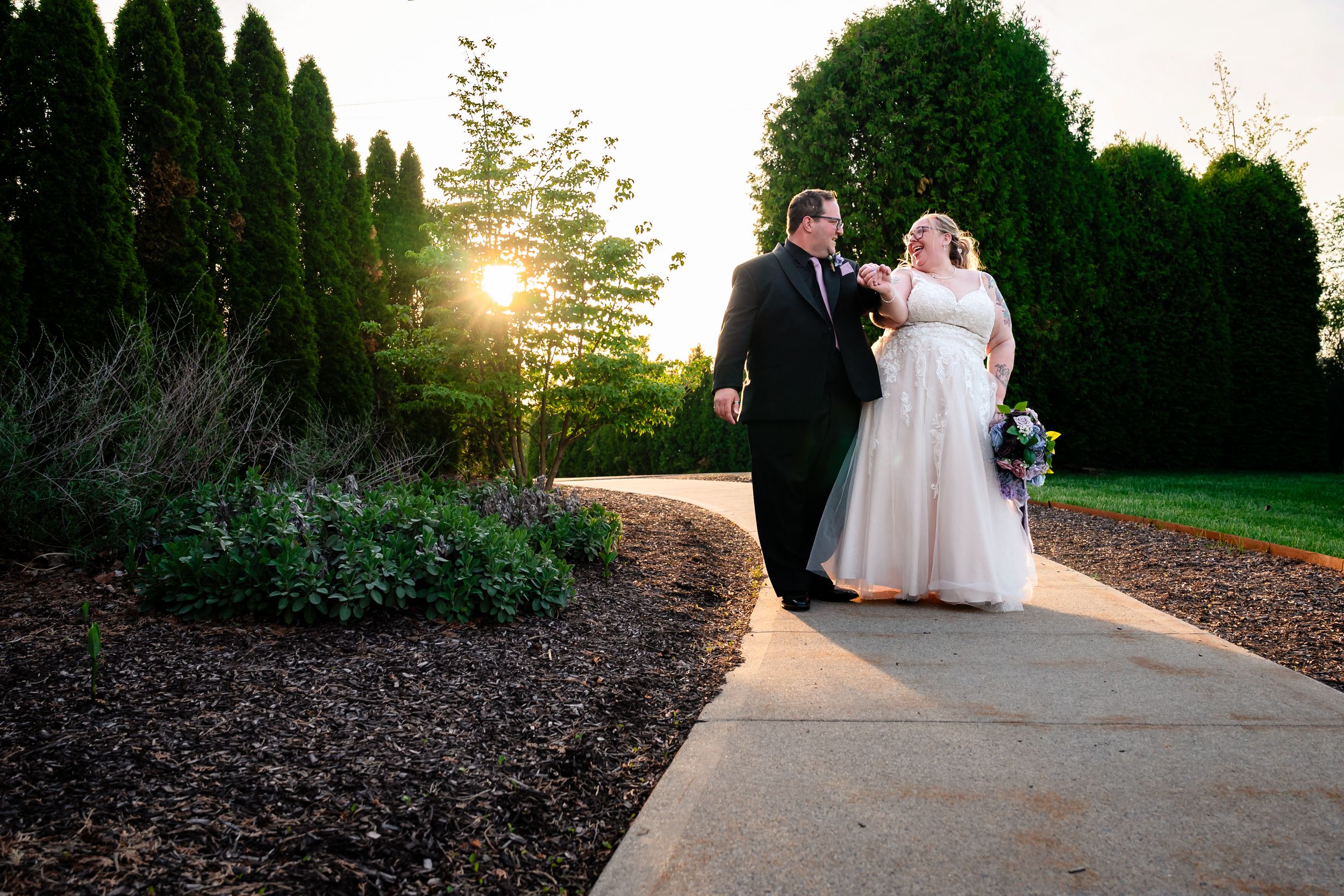 Bride and groom walking along garden path at golden hour sunset.