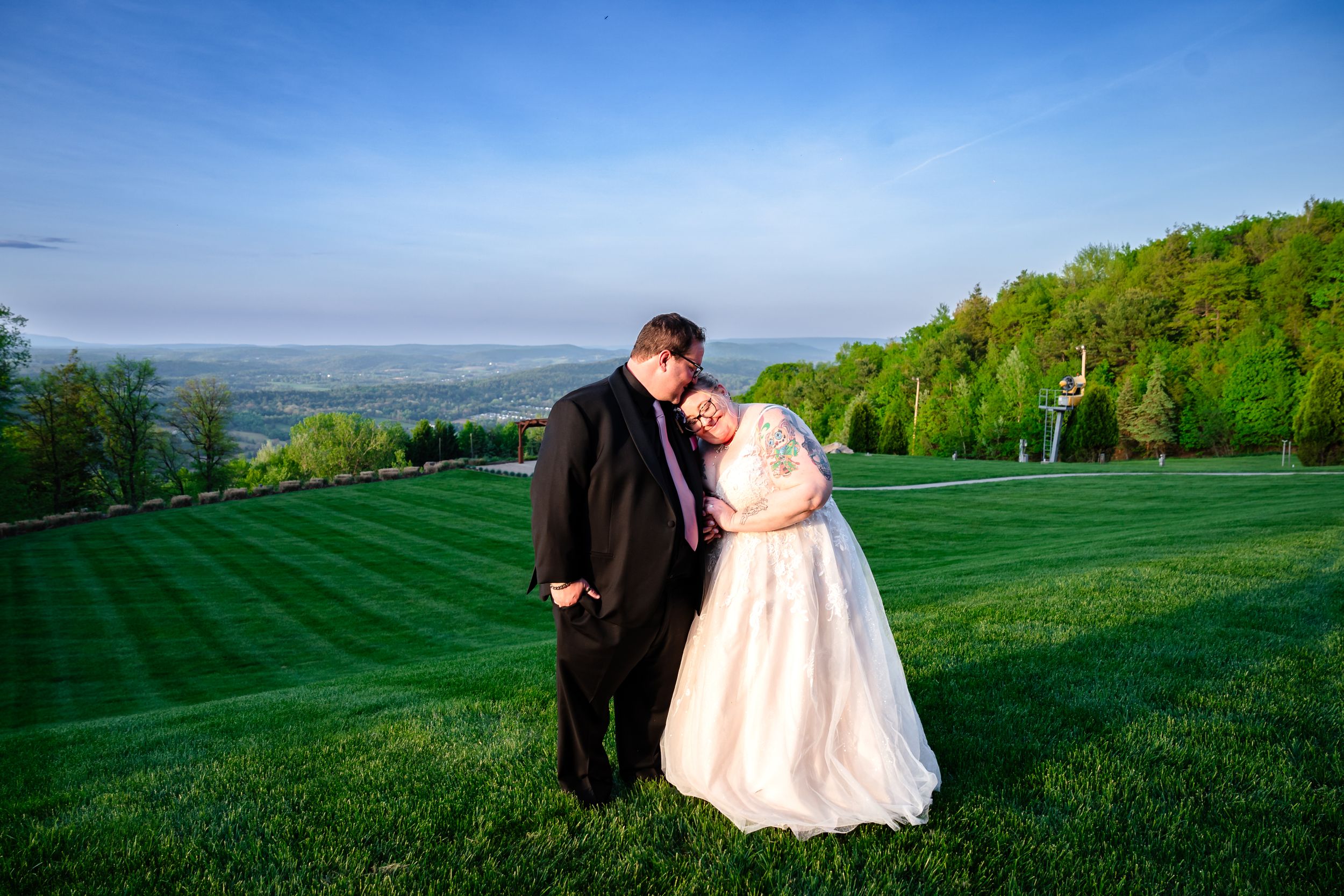 A bride and groom share a tender moment on a lush green hillside with a scenic valley view at sunset.