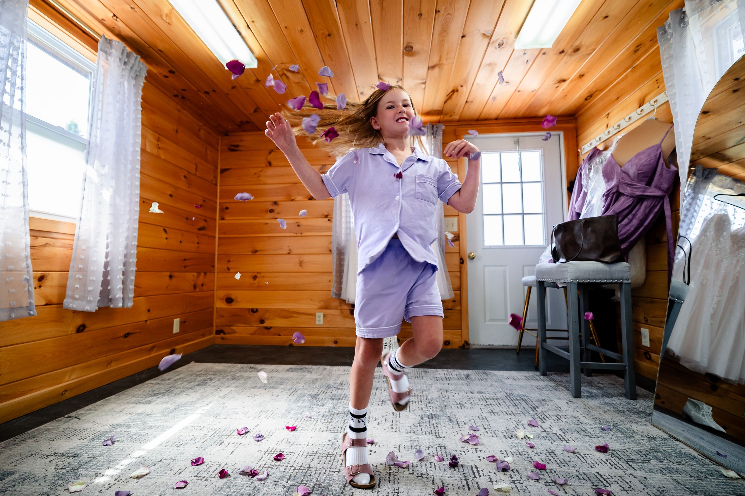 Joyful girl in purple outfit dancing indoors in a wood-paneled room with colorful flower petals falling around her.