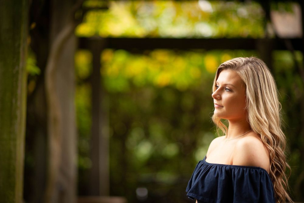 Young blonde woman in off-shoulder navy dress gazing thoughtfully in a lush green garden setting.