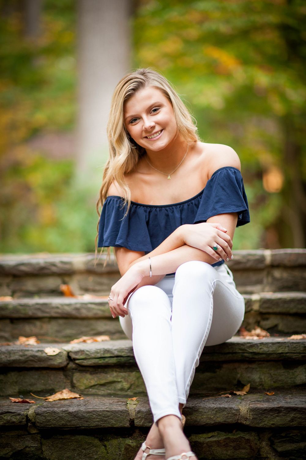 Smiling blonde teen girl in navy off-shoulder top and white jeans sitting on stone steps outdoors in autumn.