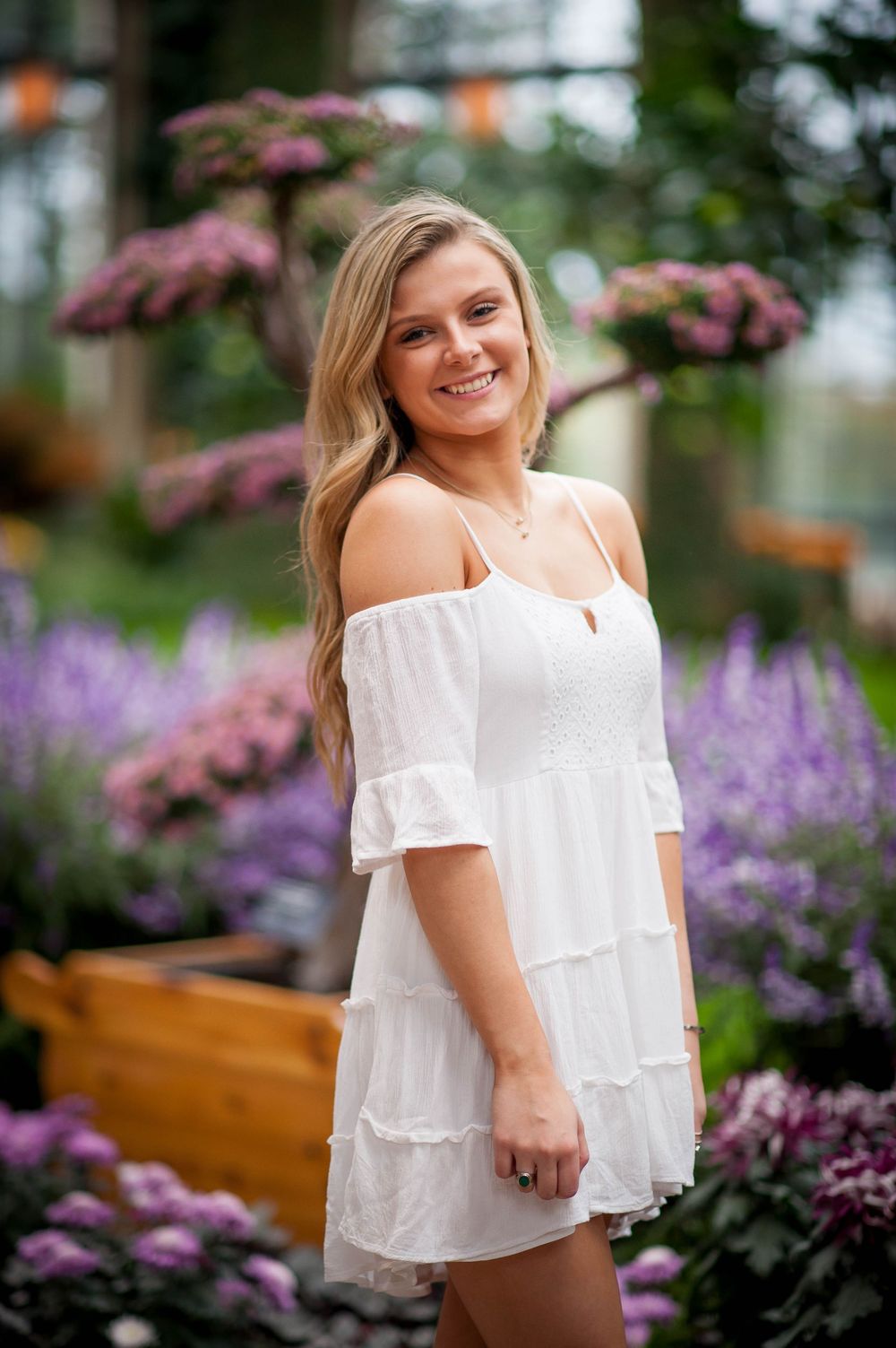 Young woman in white off-shoulder dress smiling among purple flowers in a garden greenhouse.