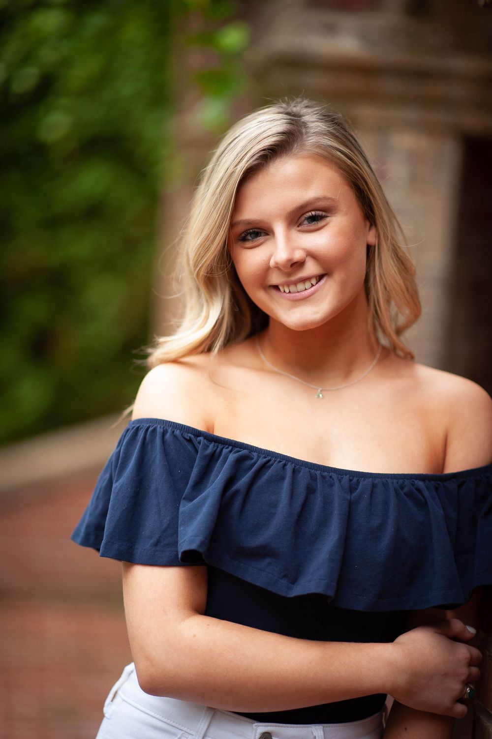 Young blonde woman smiling in navy off-shoulder top, posing outdoors near brick wall with green foliage.