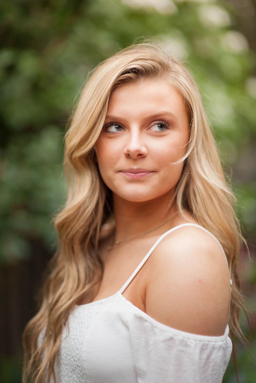 Young blonde woman in white off-shoulder top poses outdoors with green foliage background.