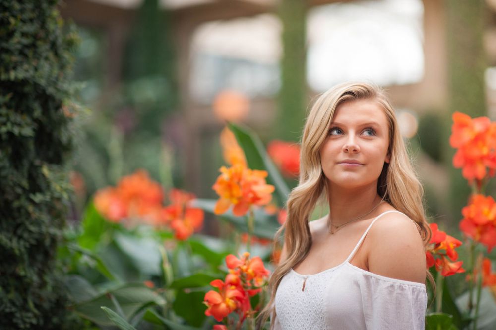 Young blonde woman in white off-shoulder top surrounded by vibrant orange tropical flowers in a lush garden setting.