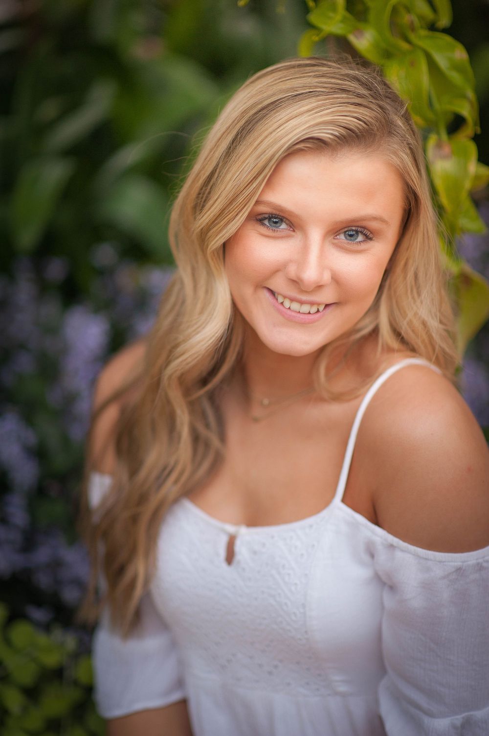 Smiling teenage girl with blonde hair and blue eyes wearing a white off-shoulder dress posed in front of lush greenery.