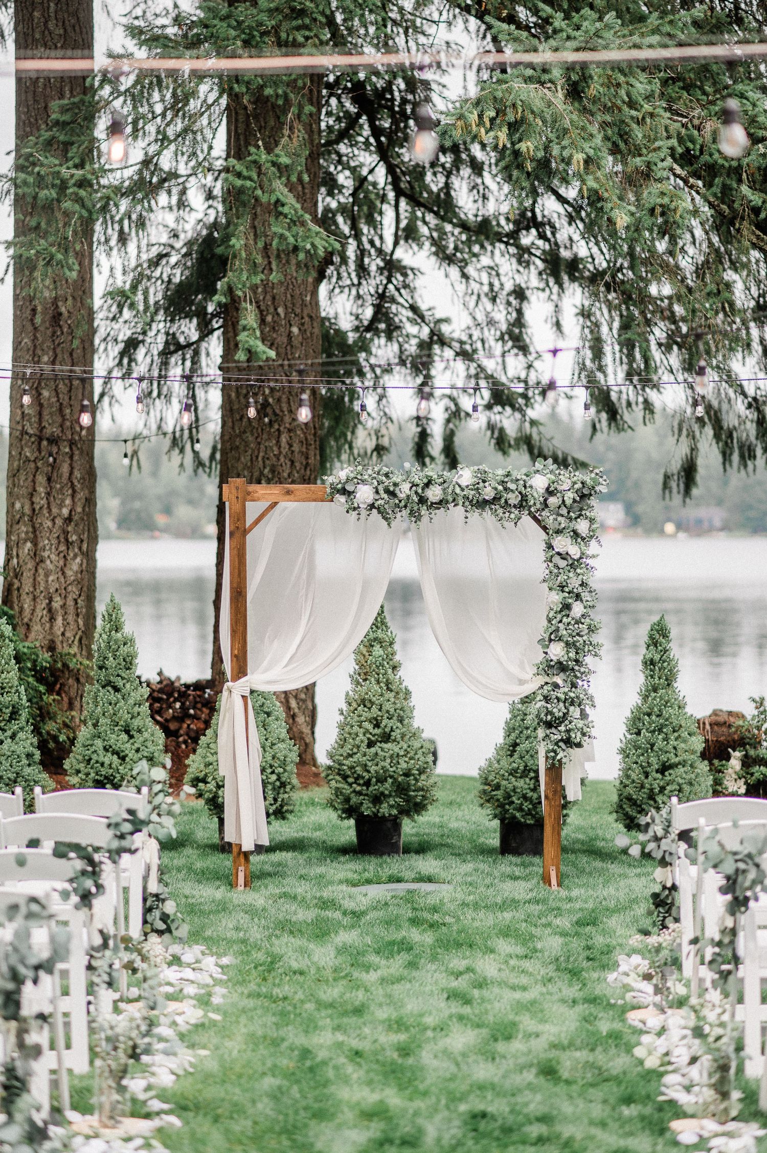 Elegant outdoor wedding ceremony arch draped in white fabric with greenery, flanked by topiary trees beside a serene lake in Bremerton, WA