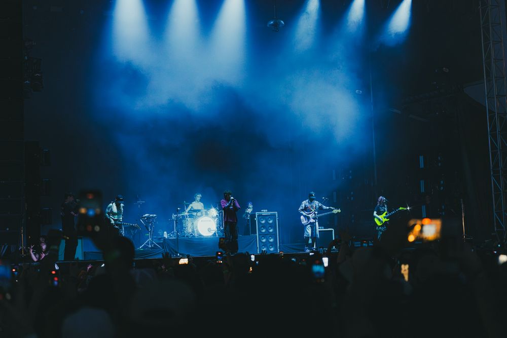 Band performing on a smoky blue-lit concert stage with crowd silhouettes in the foreground.