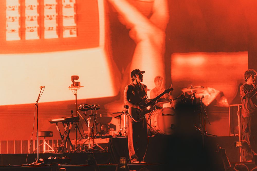 Band performing on stage bathed in red light with large projection screen backdrop at live concert.