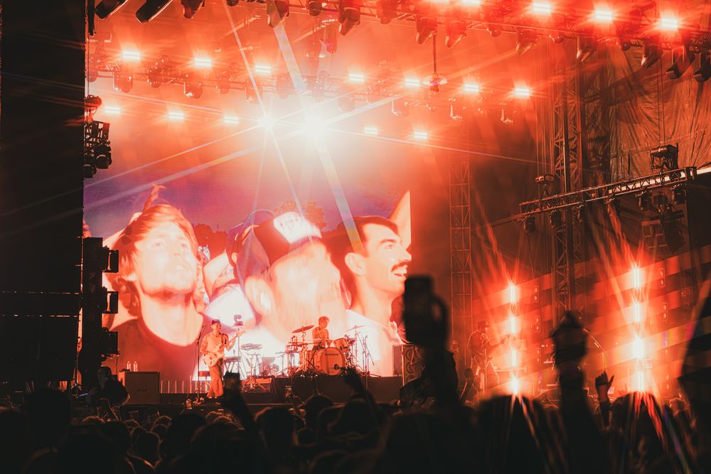 Crowd at outdoor rock concert with red stage lights illuminating performers on large screen at night.