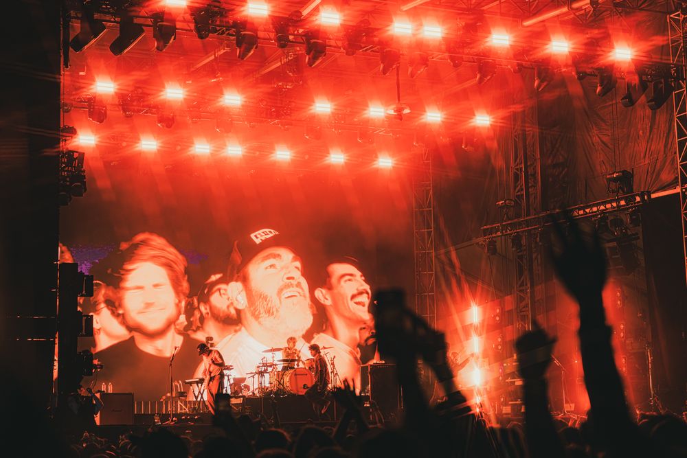Concert stage with red lights illuminating a large crowd and band members projected on a massive screen.