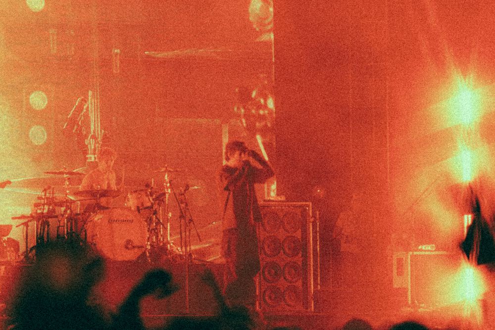 Rock band performing on a red-lit stage with crowd silhouettes and dramatic lighting effects.