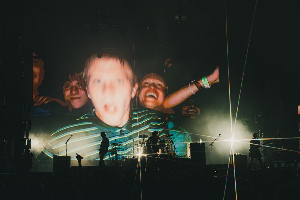 Concert crowd projected on stage screen with silhouetted performers and bright stage lights at a live music event.