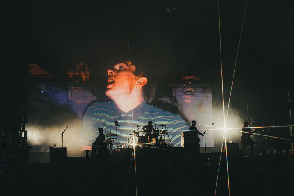 Band performing on stage with dramatic large-scale projected face backdrop and atmospheric lighting at a concert.