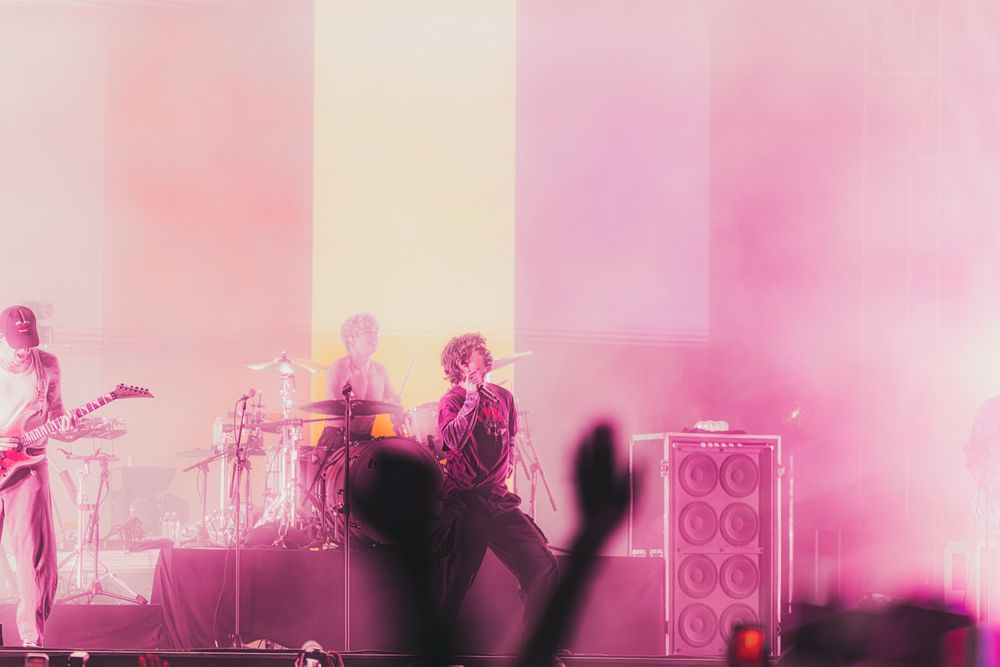 Band performing on stage bathed in pink and red lights with amplifiers and silhouetted crowd in foreground.