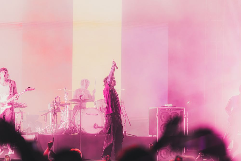 A band performs on stage bathed in pink and yellow lights with a singer raising their arm to the crowd.