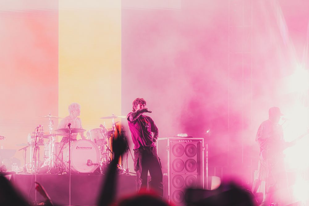 A singer performs on a foggy pink-lit concert stage with band members and speaker stacks visible in the background.
