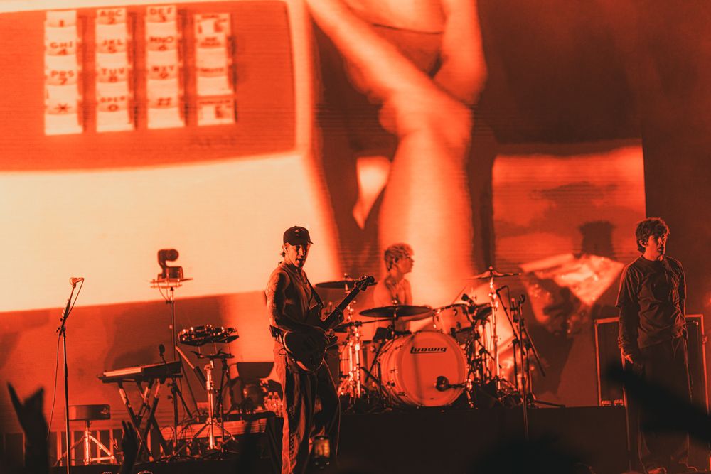 Three musicians perform on a red-lit stage with drums and keyboards, large projection screen behind them.