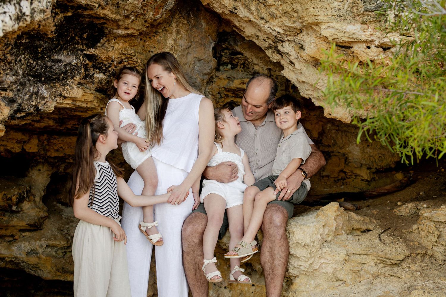 Family of six posing together in a natural rock cave, parents with four children dressed in white and neutral tones.