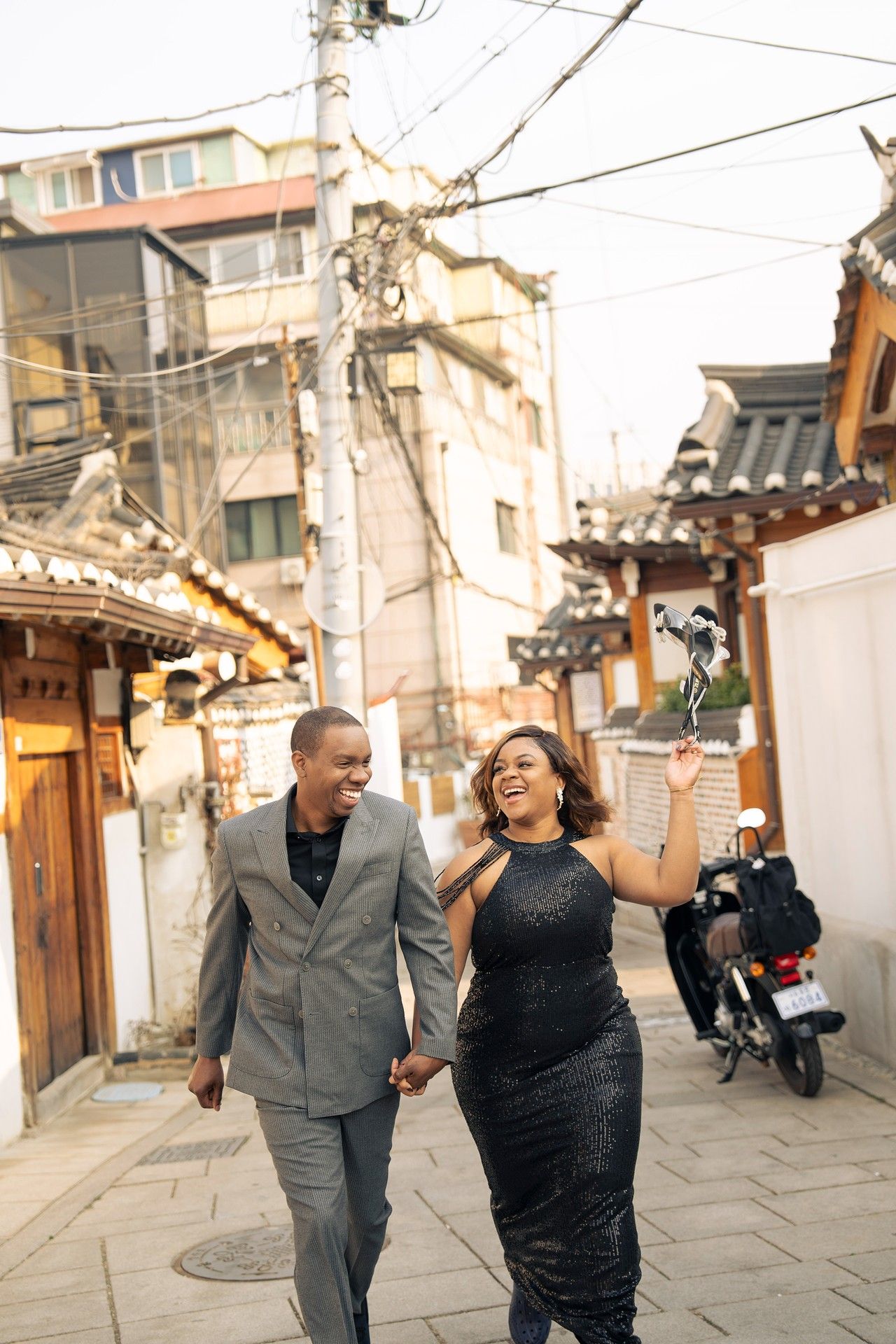 A joyful Black couple walks hand-in-hand through a traditional Korean alley, dressed in elegant formal attire.