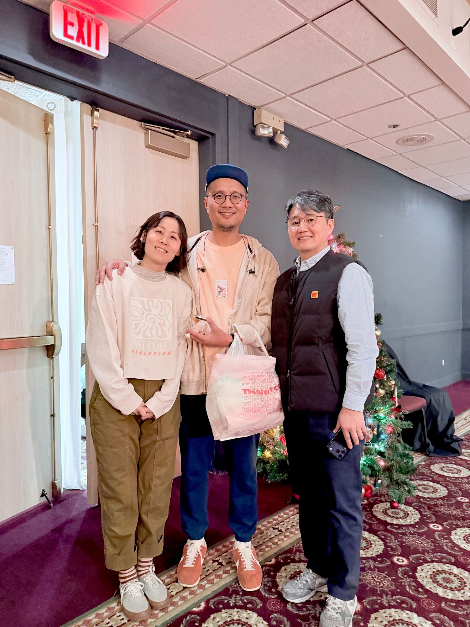 Three people smiling together near a Christmas tree and exit sign at an indoor holiday gathering.