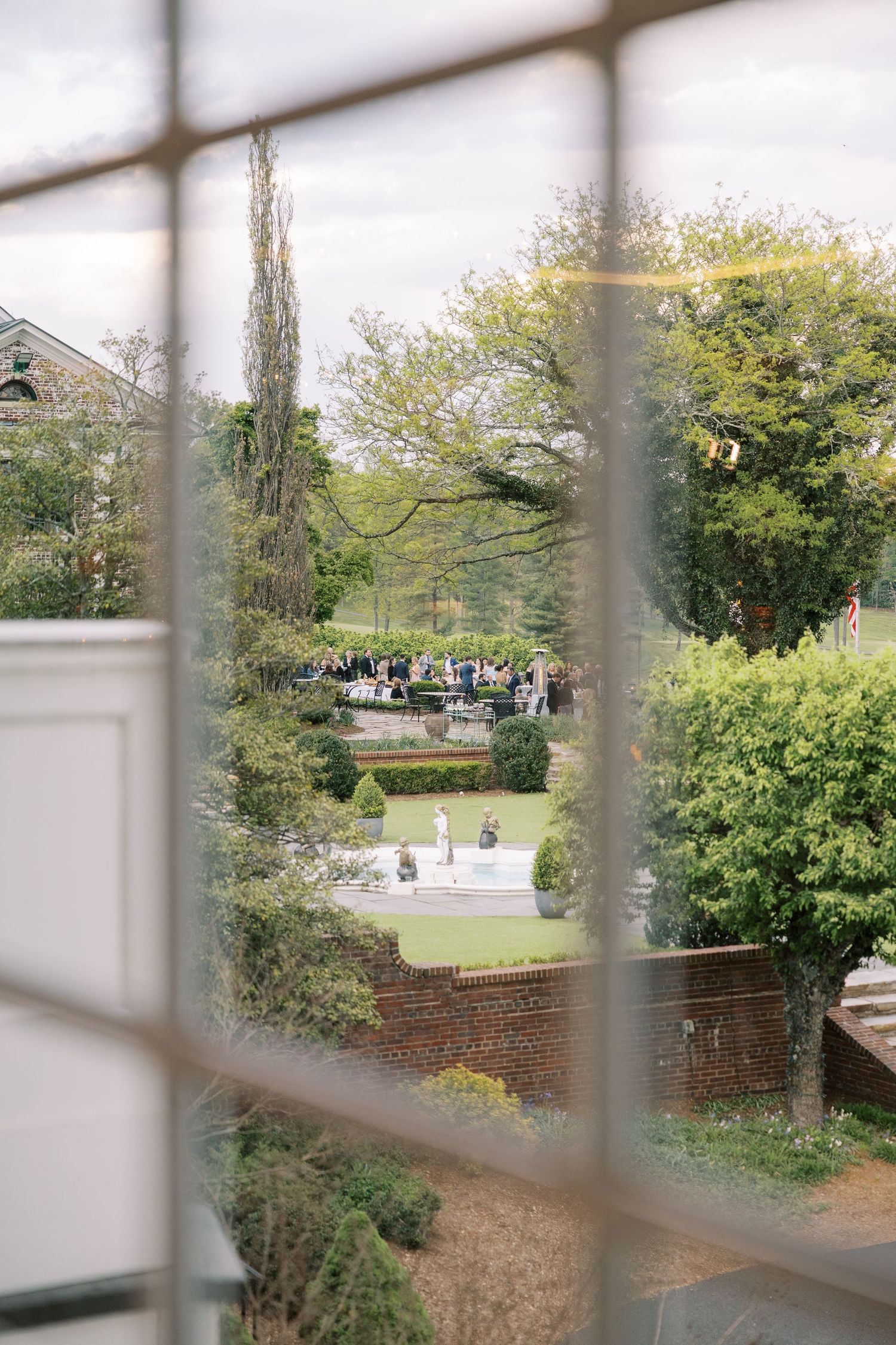 View through a window of an elegant garden wedding ceremony surrounded by lush greenery and manicured hedges.