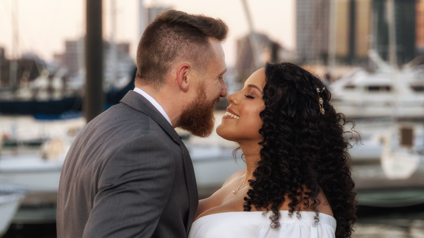 Couple sharing romantic moment at waterfront marina, bride in white dress and groom in gray suit.