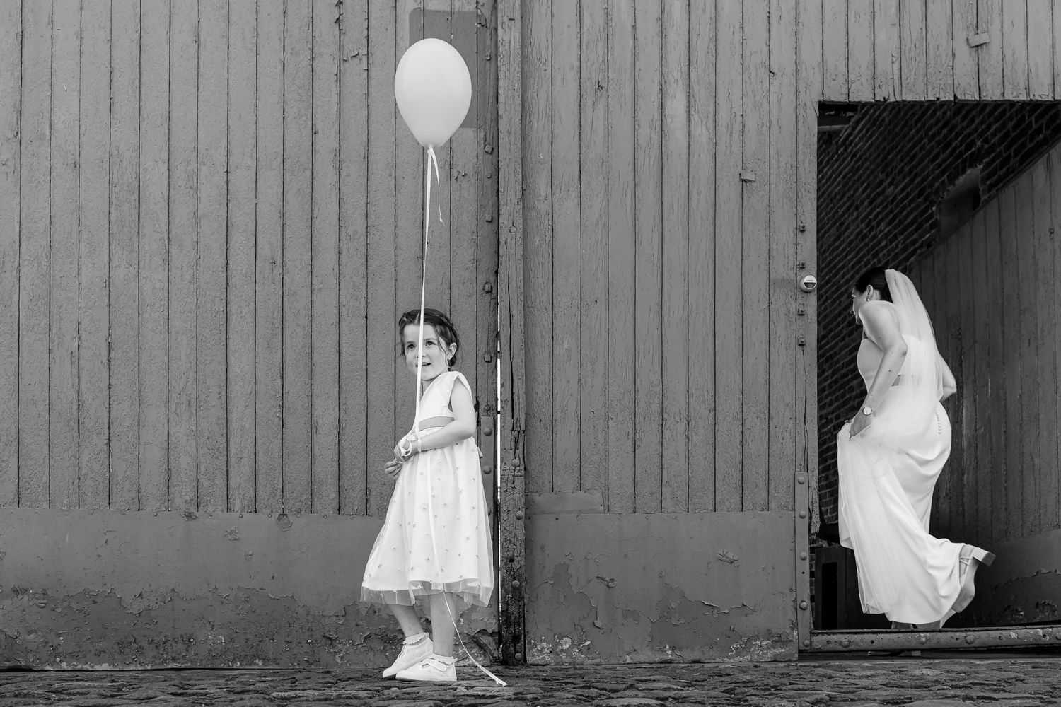 Black and white wedding photo of flower girl holding balloon beside barn as bride enters doorway.