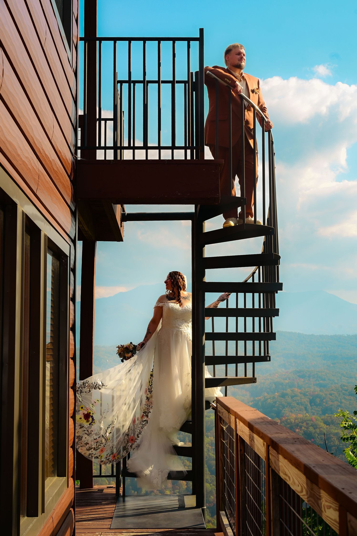 Bride and groom pose on a spiral staircase of a mountain cabin, with scenic autumn forest views under blue skies.