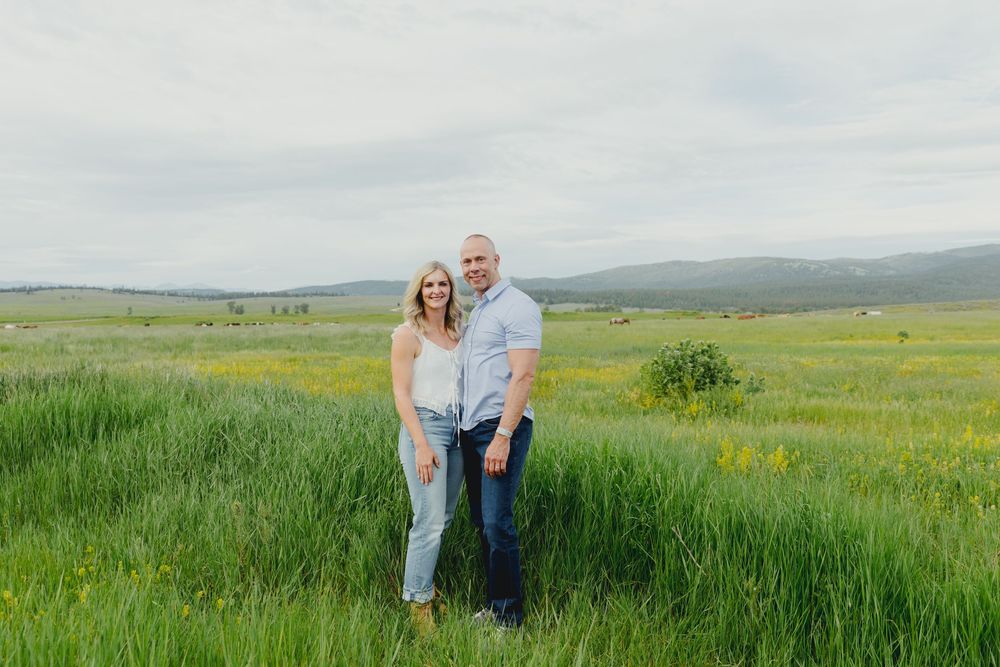 Couple smiling together in a lush green meadow with yellow wildflowers and rolling hills under a cloudy sky.