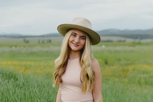 Young blonde woman wearing a tan wide-brim hat smiling in a green meadow with mountains behind her.