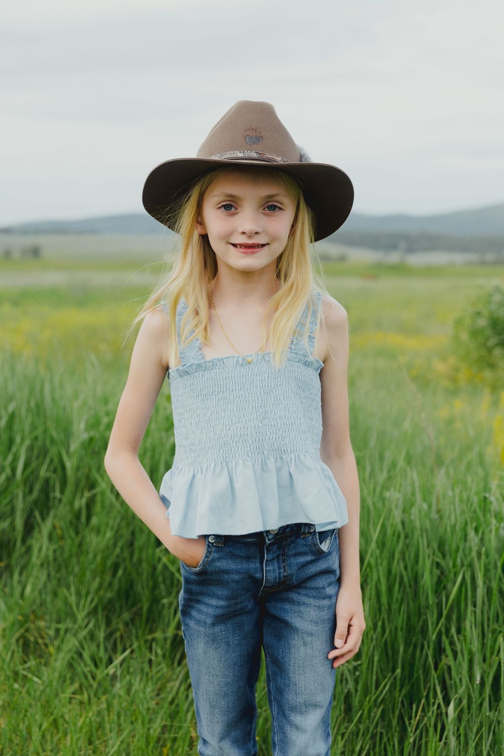 A smiling blonde girl wearing a brown cowboy hat, denim top, and jeans standing in a green field.