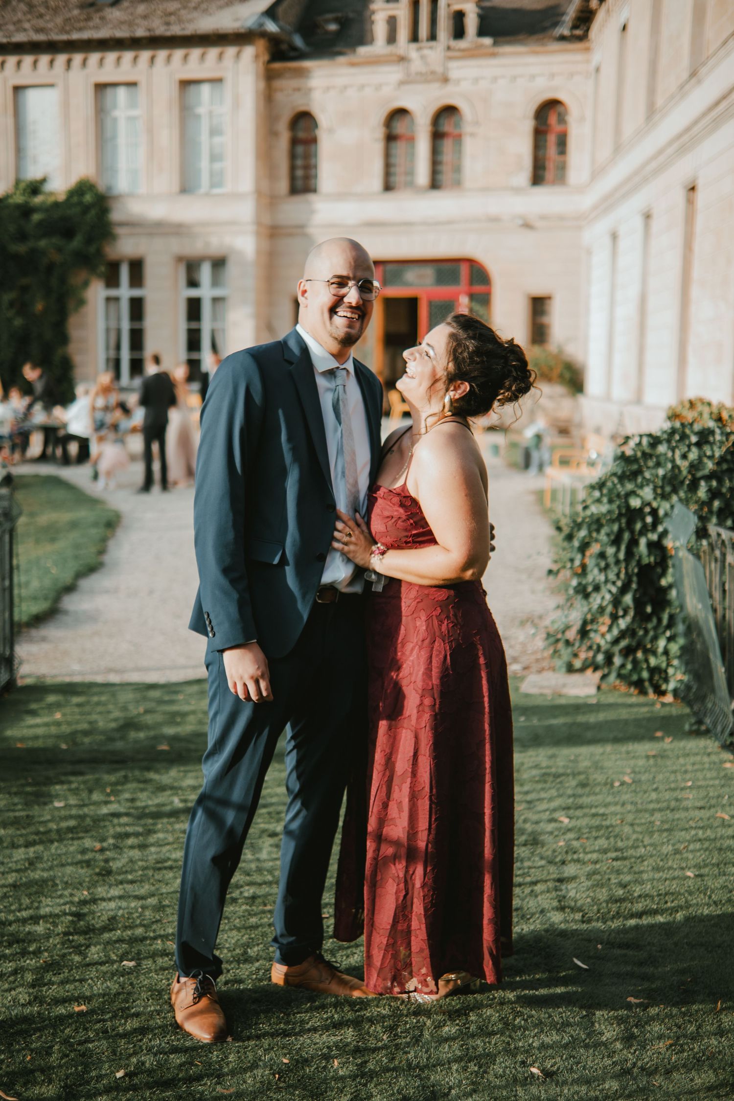 Couple smiling and embracing outdoors at a wedding venue, man in navy suit, woman in burgundy lace gown.