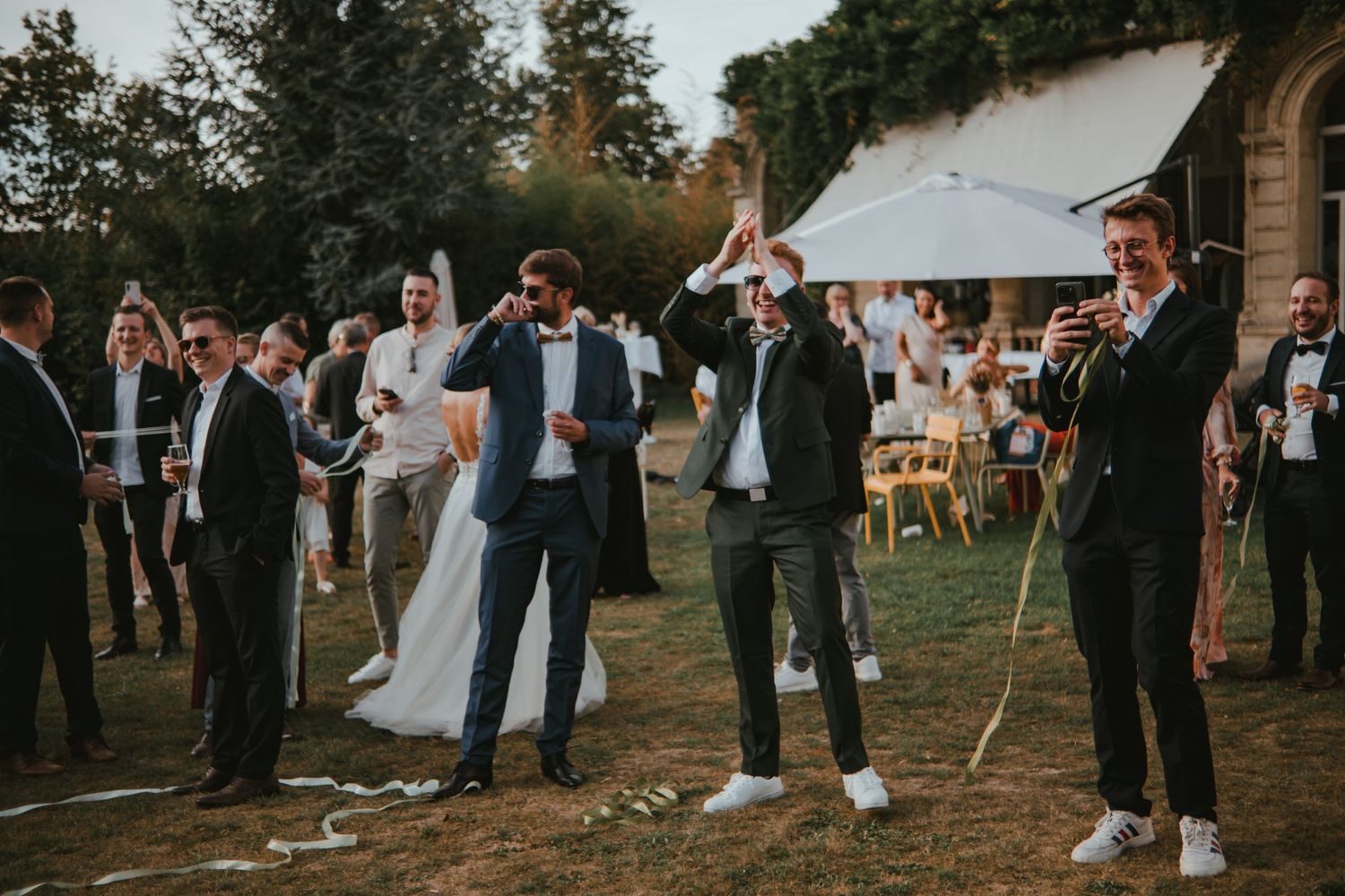 Wedding celebration outdoors with guests cheering, bride and groom dancing, and a trumpeter performing on a sunny day.