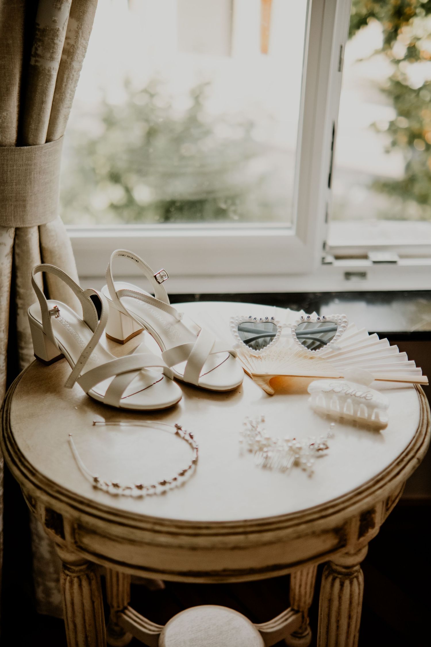 White bridal heels, pearl jewelry, heart sunglasses, and a fan arranged on a vintage table by a window.