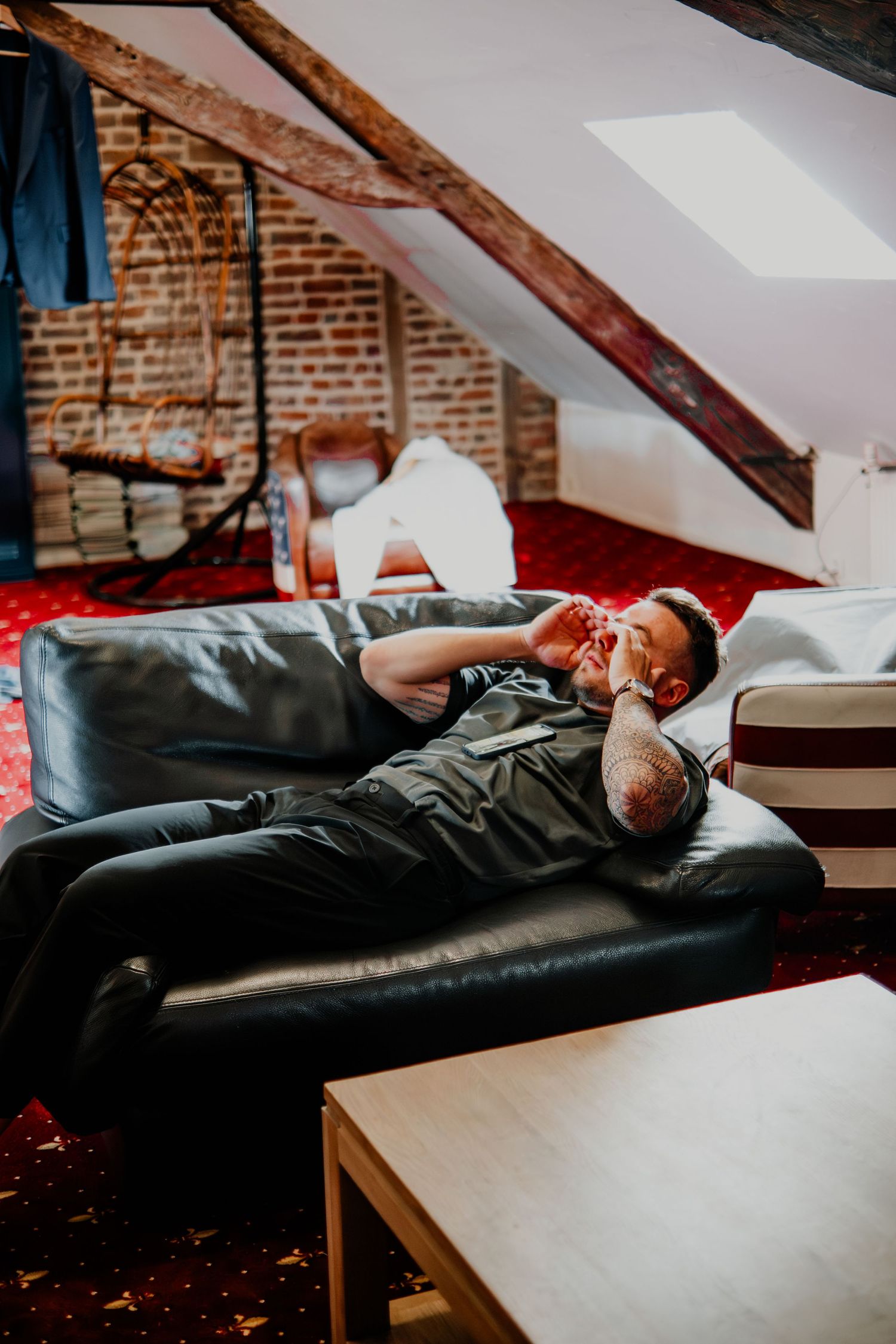 Man relaxing on a black leather sofa in a rustic attic room with exposed brick walls and wooden beams.