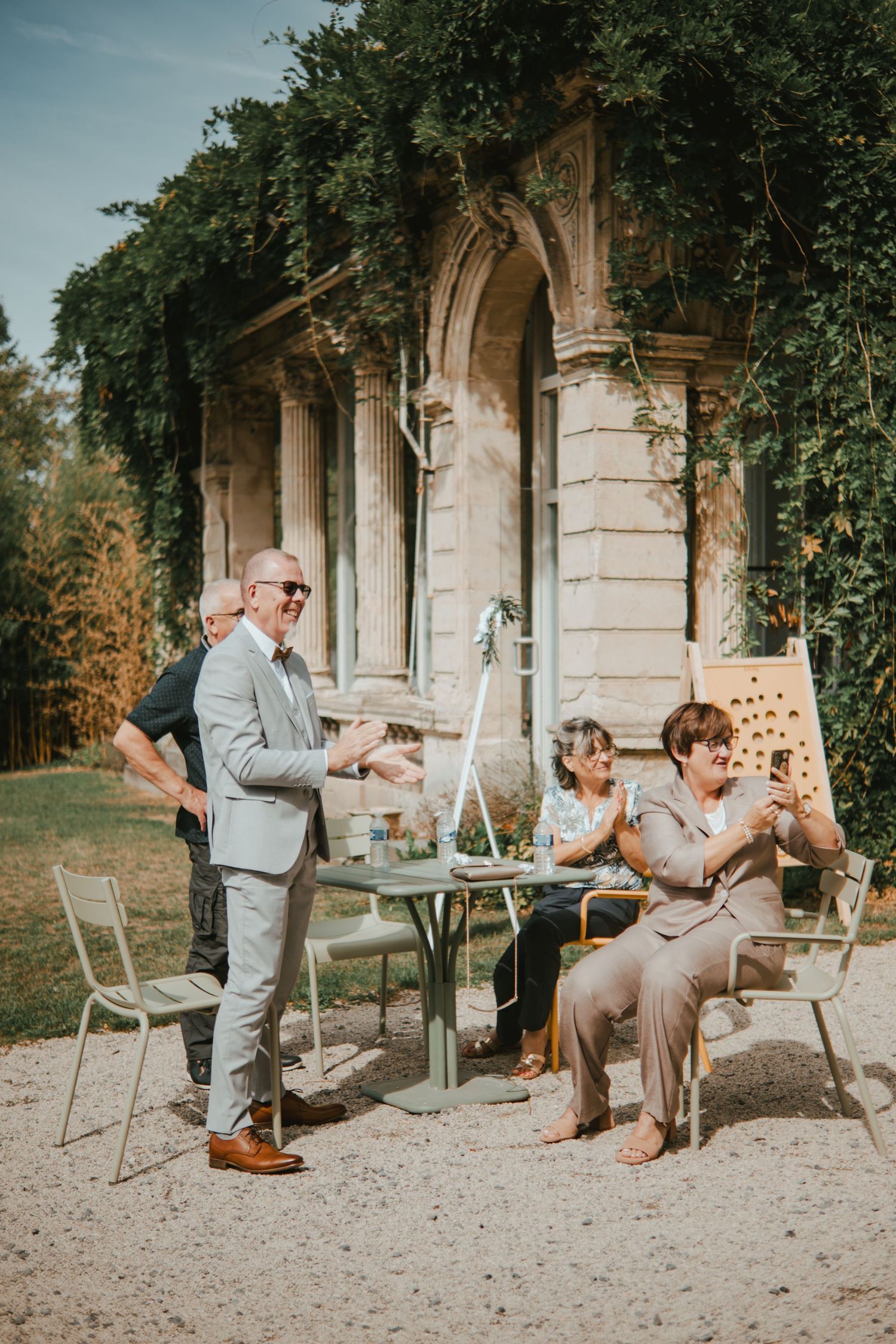 Elegant outdoor gathering at a historic stone pavilion with ivy, guests in formal attire clapping and socializing.