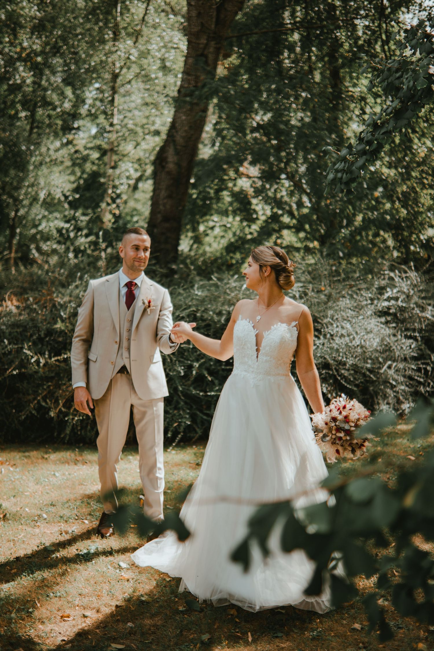 Bride and groom hold hands in a lush forest setting, she in a white lace gown, he in a beige suit.
