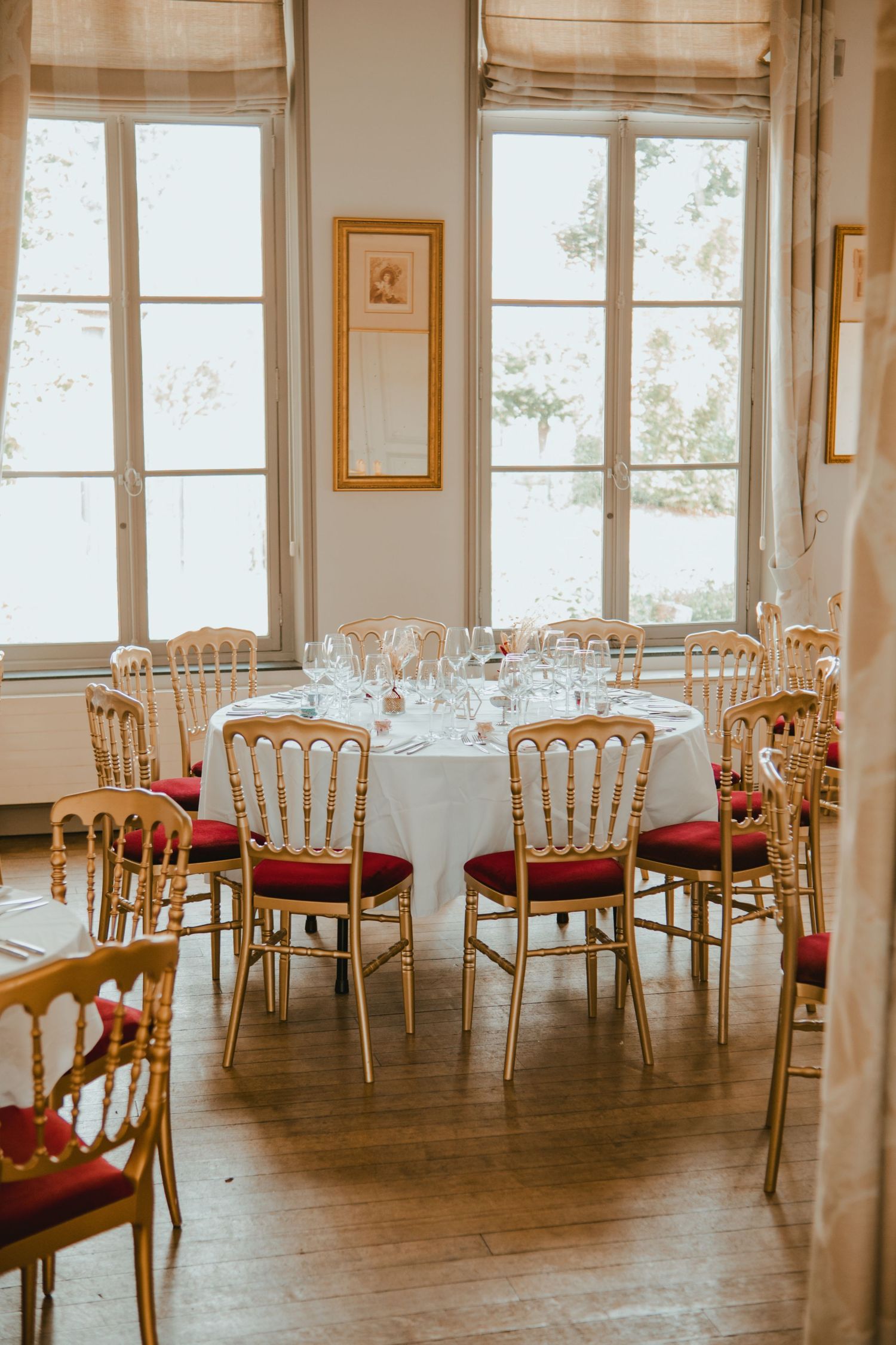 Elegant dining room with gold Chiavari chairs, red cushions, and round tables set with glassware near bright French windows.