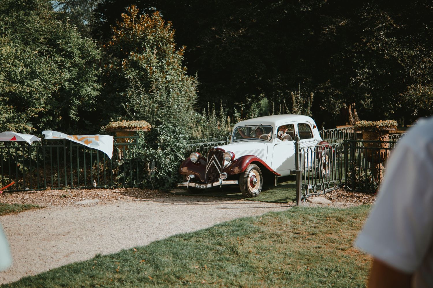 Vintage white and red Citroën Traction Avant parked in a lush garden setting with iron gates and trees.