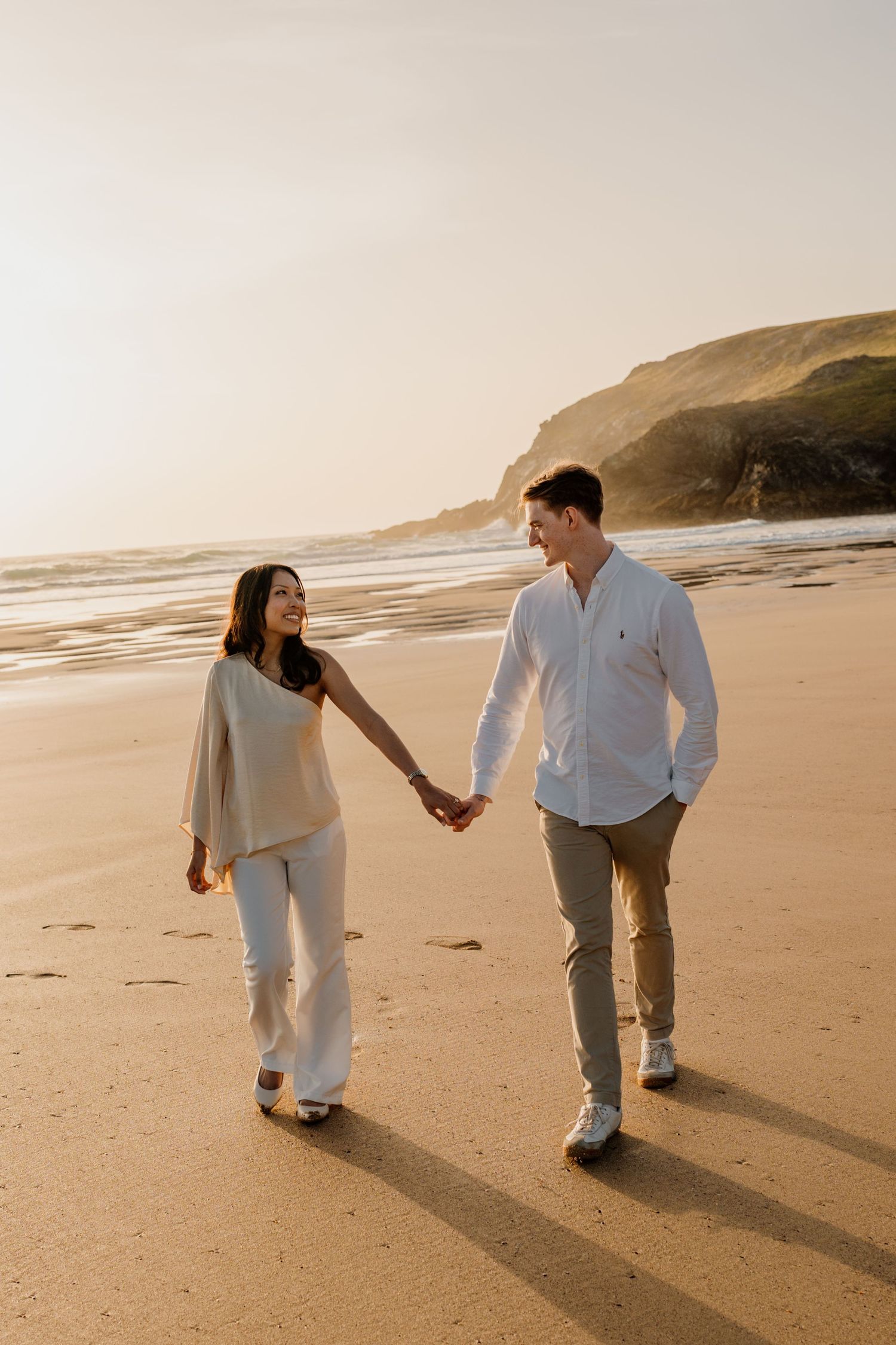 Couple holding hands and walking on a sandy beach at golden hour with cliffs in the background.