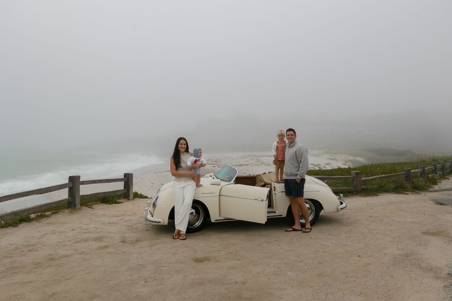 Couple with dog posing beside vintage white convertible car on foggy coastal overlook with ocean views.