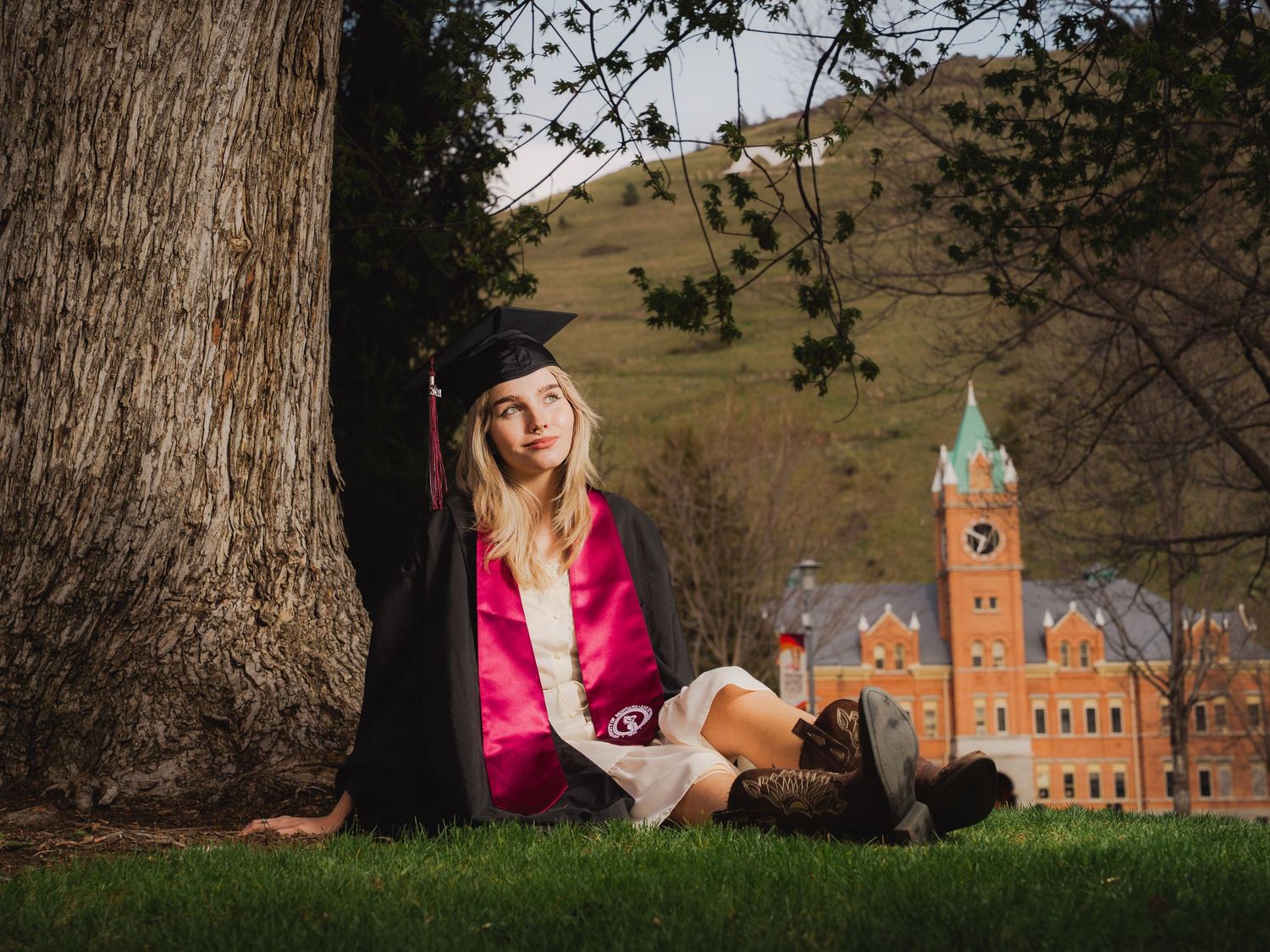 Graduate in cap and gown sitting by a tree on green grass with a historic university building in the background.