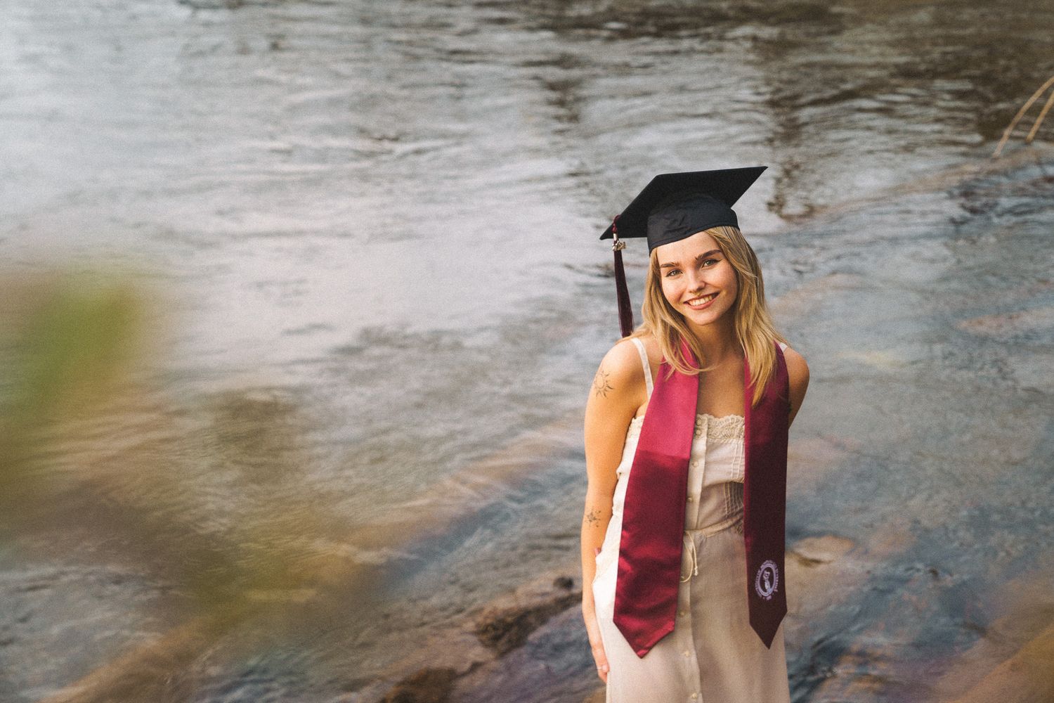 Smiling female graduate in cap and stole posing by a rocky creek in an outdoor graduation portrait.