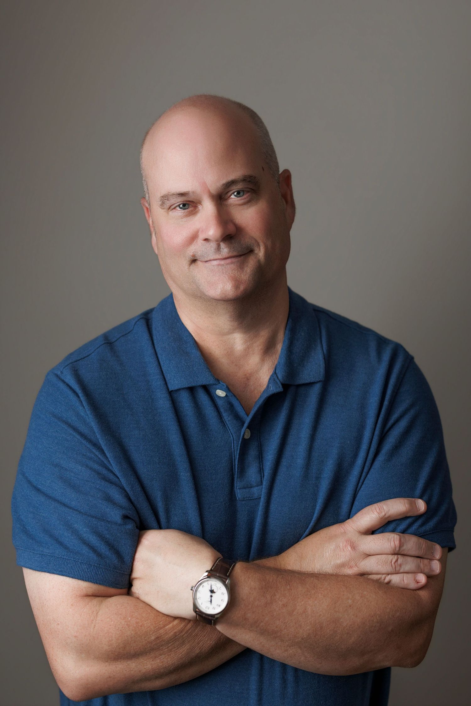 Bald middle-aged man in blue polo shirt with arms crossed, smiling confidently, wearing a watch, studio portrait.